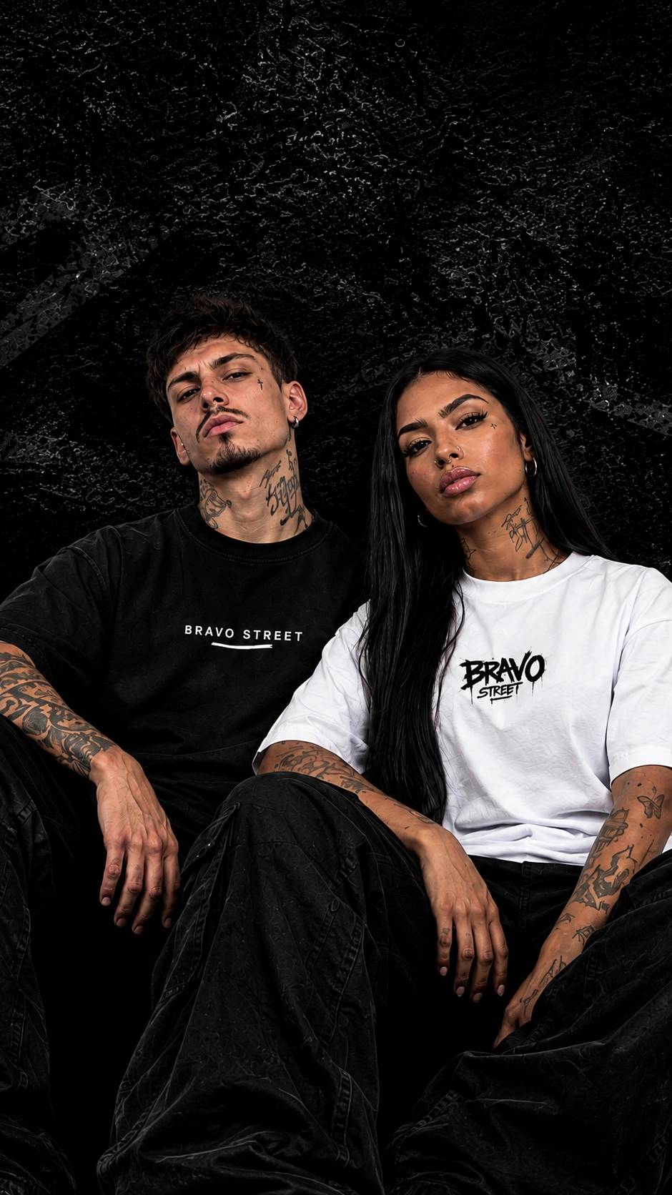 Um homem de camiseta preta Bravo Street e uma mulher de camiseta branca Bravo Street sentados diante de um muro escuro